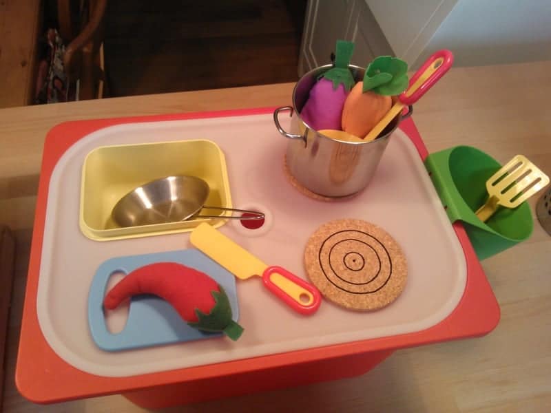 Toy kitchen playset on a table with a metal pot of felt vegetables and various plastic utensils and accessories for pretend cooking.
