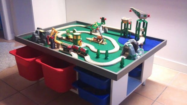 Miniature wooden train set on a raised play table with red and blue storage bins underneath.