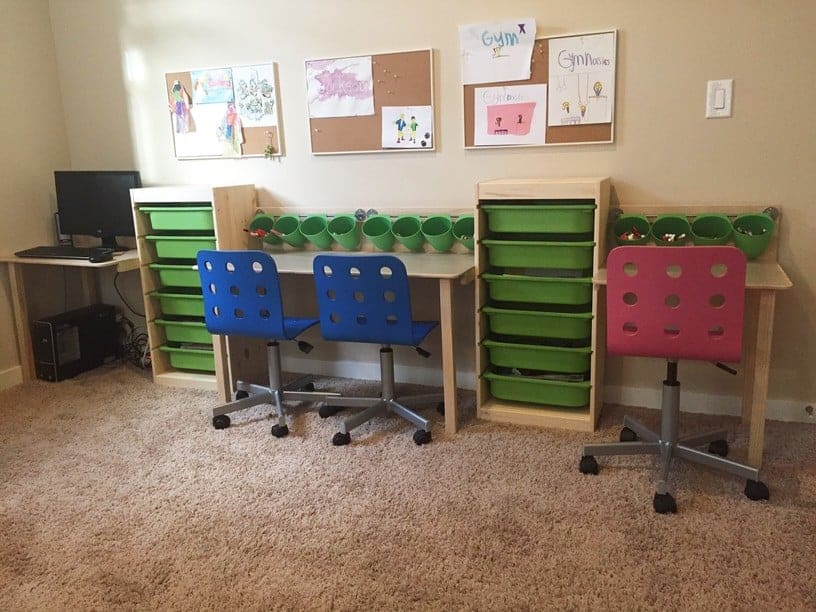 A classroom study area with two blue rolling chairs at a wooden desk, green storage drawers, and a pink chair by the wall.