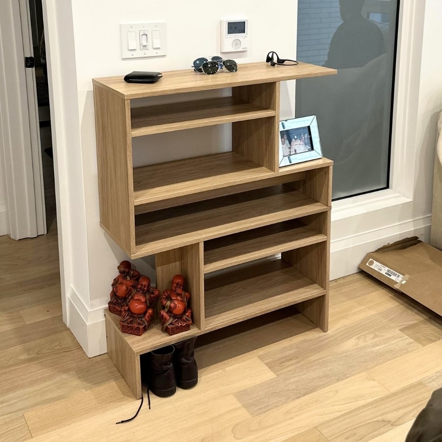 IKEA HOLMERUD Light wood multi-tier shelving unit with five open compartments placed against a wall near a window; top has sunglasses and a small device, with a framed photo on one shelf and two red Buddha statues nearby.