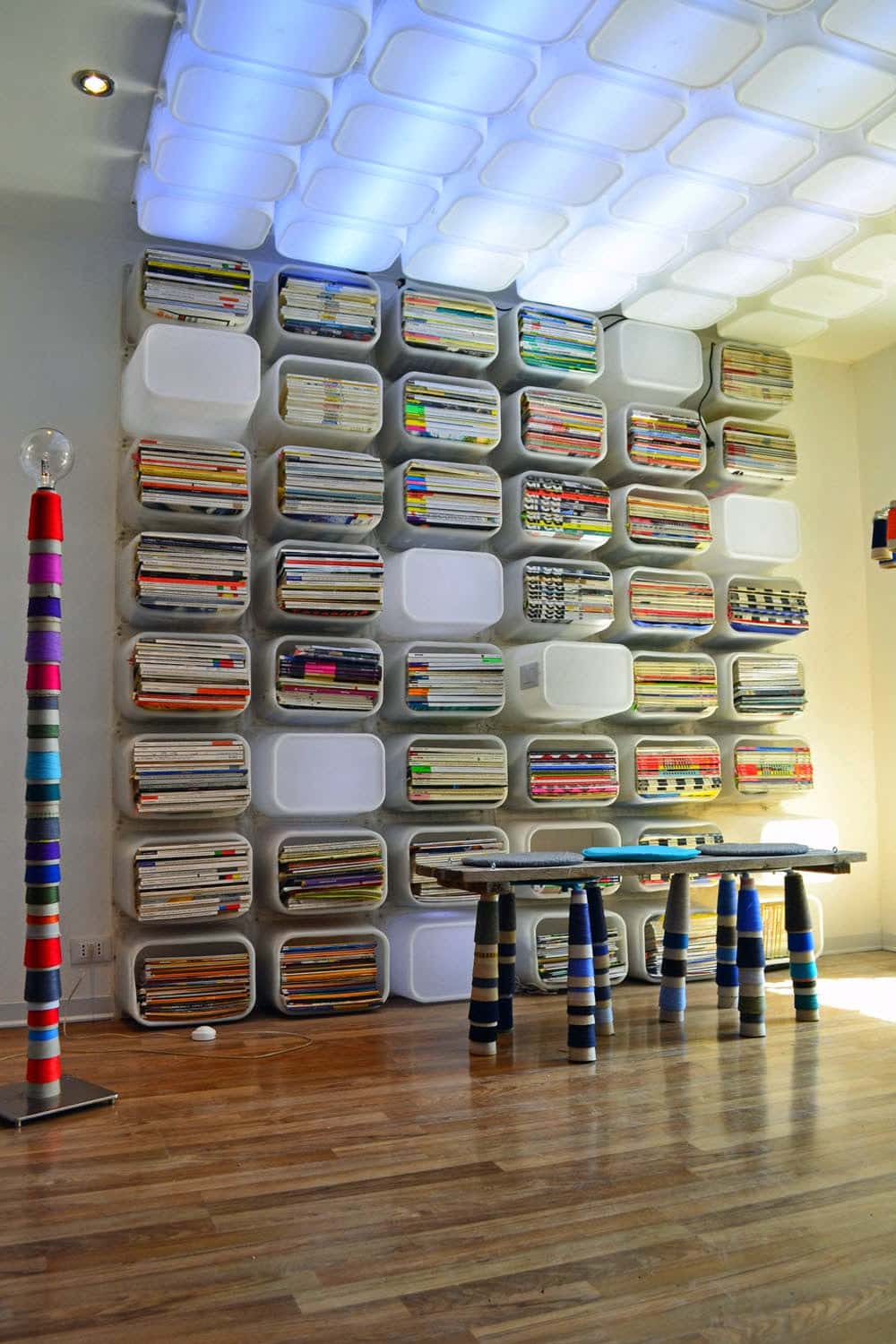 White modular shelves filled with colorful stacked magazines arrange a geometric wall installation; a striped column and striped-taped table legs sit in a bright gallery space.