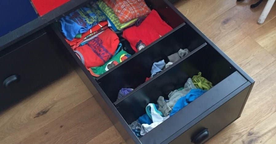 Open dresser drawer with divided compartments containing folded colorful clothes and socks, on a wooden floor.