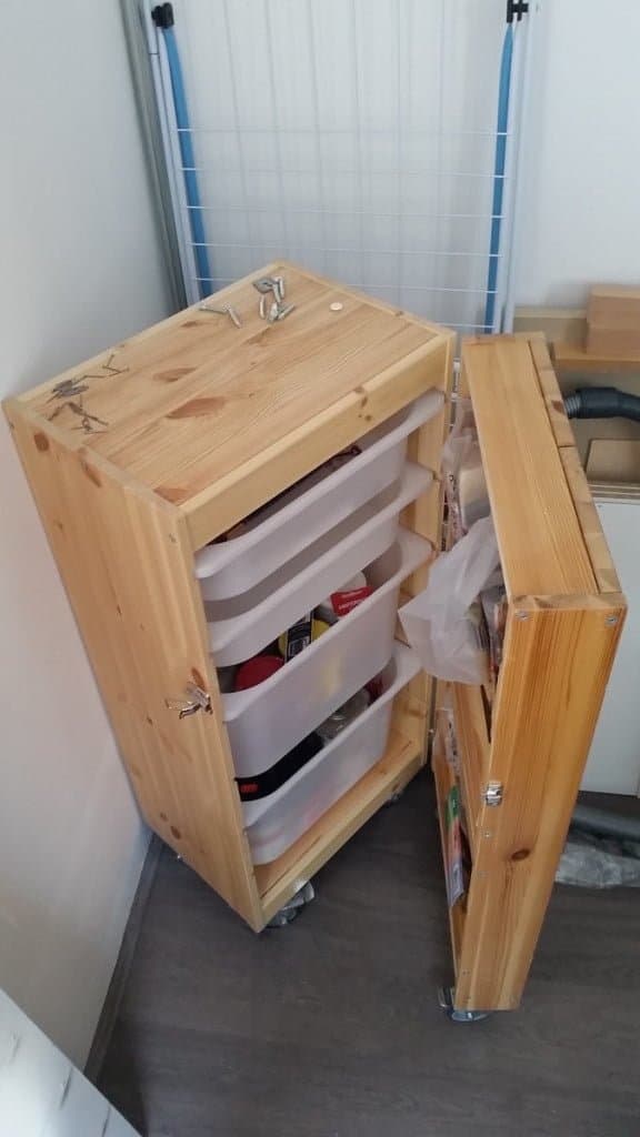 Wooden cabinet on wheels with open door, stacked white plastic bins inside stored tools and supplies in a narrow corner.
