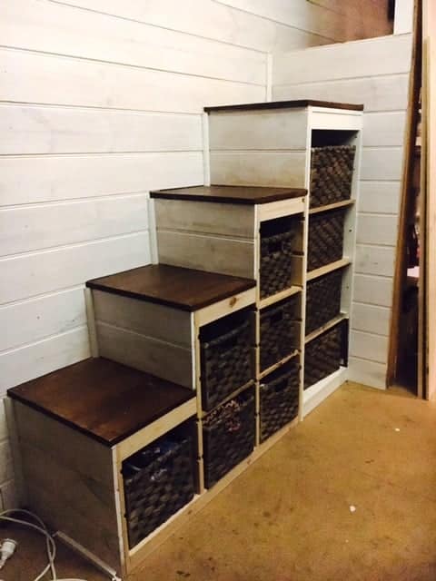 Tiered wooden step shelves with woven baskets in each cubby, white frame with dark brown tops, in a storage room.