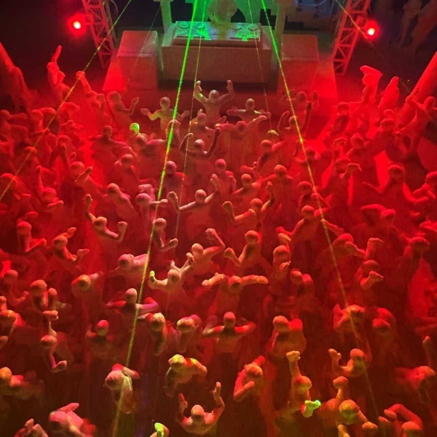 Dense crowd of small, mannequin-like figures with raised arms, illuminated in red light with green laser beams and a stage in the background.