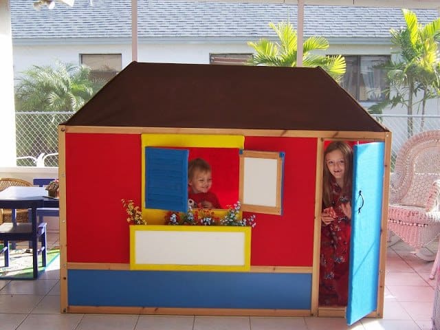 100_4317-767835 - IKEA Hackers Colorful wooden playhouse on a sunny patio with two children peeking from the windows, one at the red wall and the other near the blue door.
