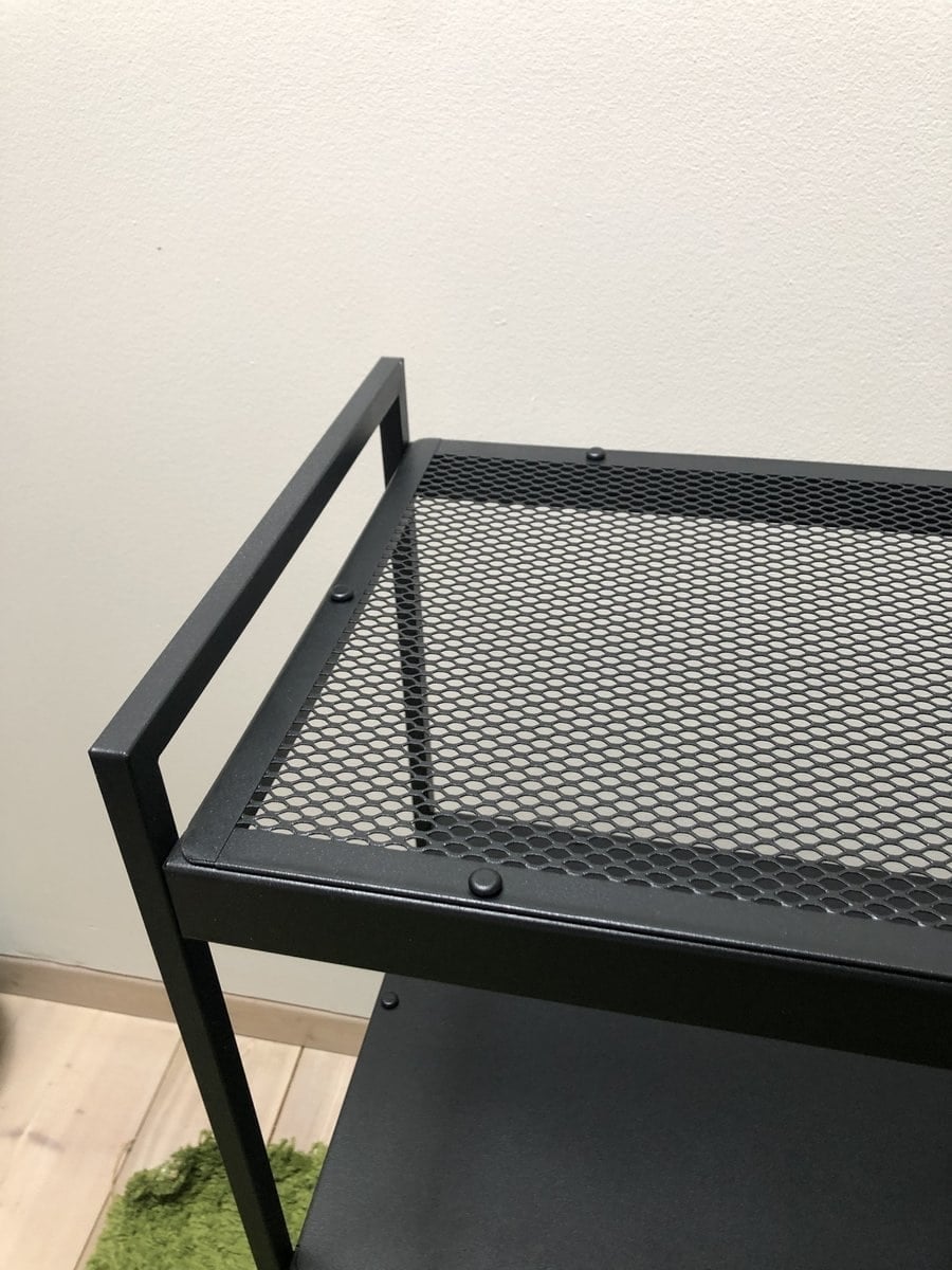 Close-up of a black metal shelving cart with a mesh top against a beige wall.