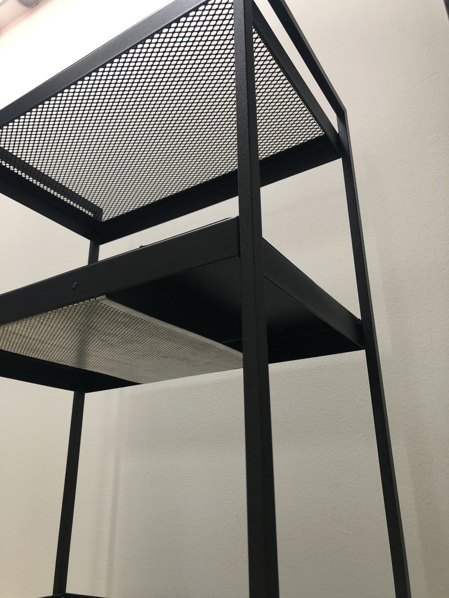 Black metal shelving unit with mesh shelves, seen from below against a pale wall.