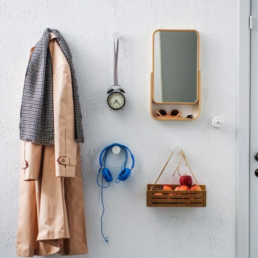 21 - IKEA Hackers Beige trench coat with a checkered scarf hangs on a wall beside a small clock, blue headphones, a wooden mirror shelf, and a crate of apples beneath it.