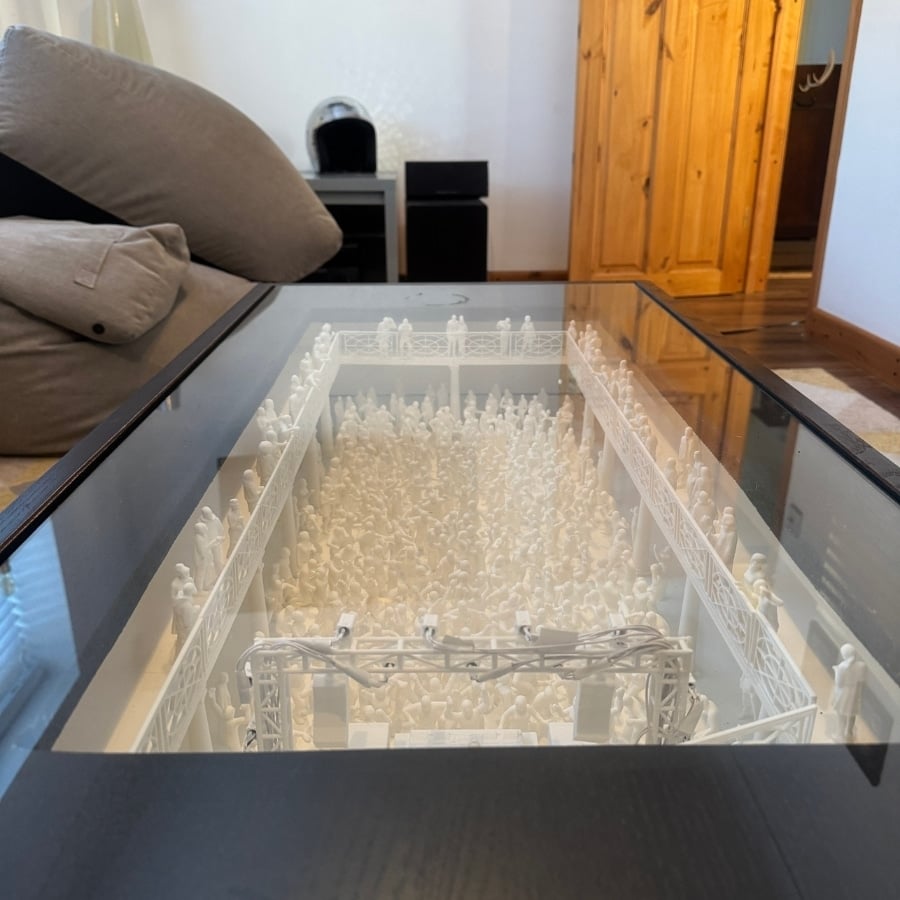 White 3D-printed crowd model inside a glass-topped table, resembling an arena with railings and scaffolding in a home setting
