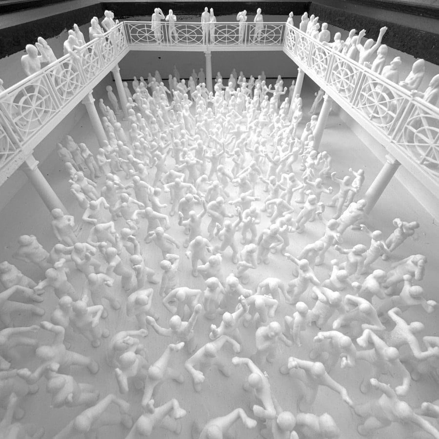Room filled with hundreds of white mannequin figures crowded in a central pit, surrounded by an ornate white balcony railing.