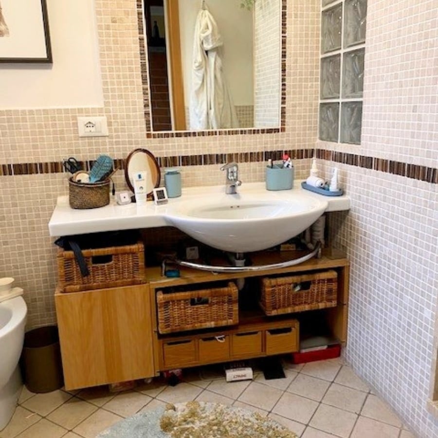 Bathroom vanity with white sink, wooden cabinet, and woven baskets for storage beneath a tiled wall mirror and small countertop items.