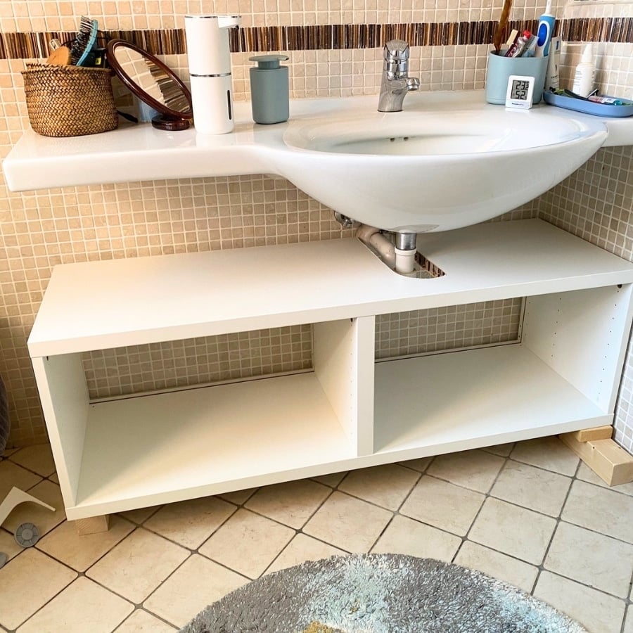 White bathroom sink on a white cabinet with open shelves beneath, against a beige tiled wall.