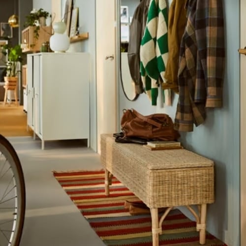 IKEA-bench-hallway - IKEA Hackers Entryway with coats hanging on wall hooks, a wicker bench with a brown bag on top, striped rug, and a bicycle wheel peeking in from the left.