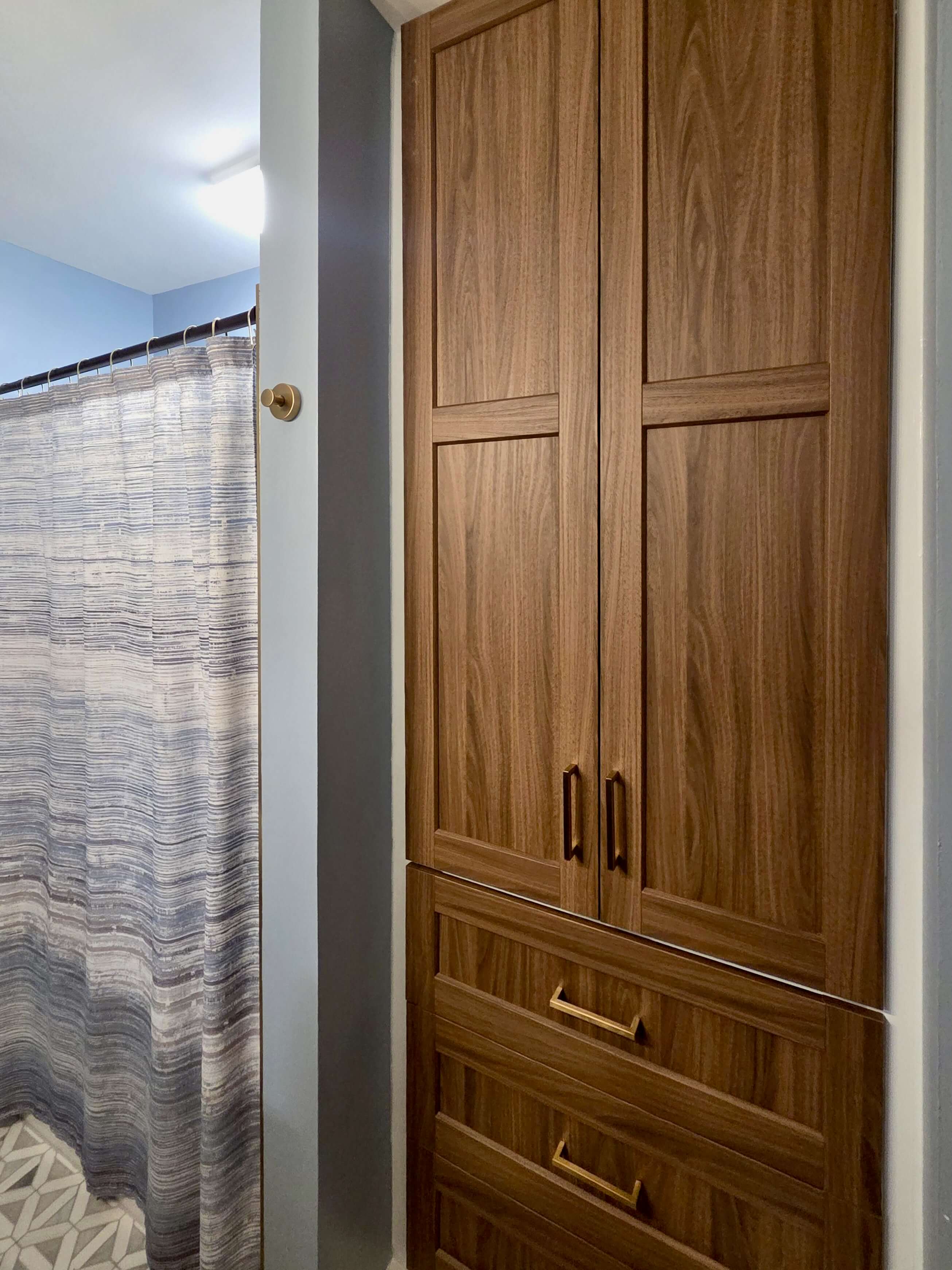 Tall wooden bathroom cabinet with two doors and three drawers, next to a striped shower curtain in a blue-gray bathroom.