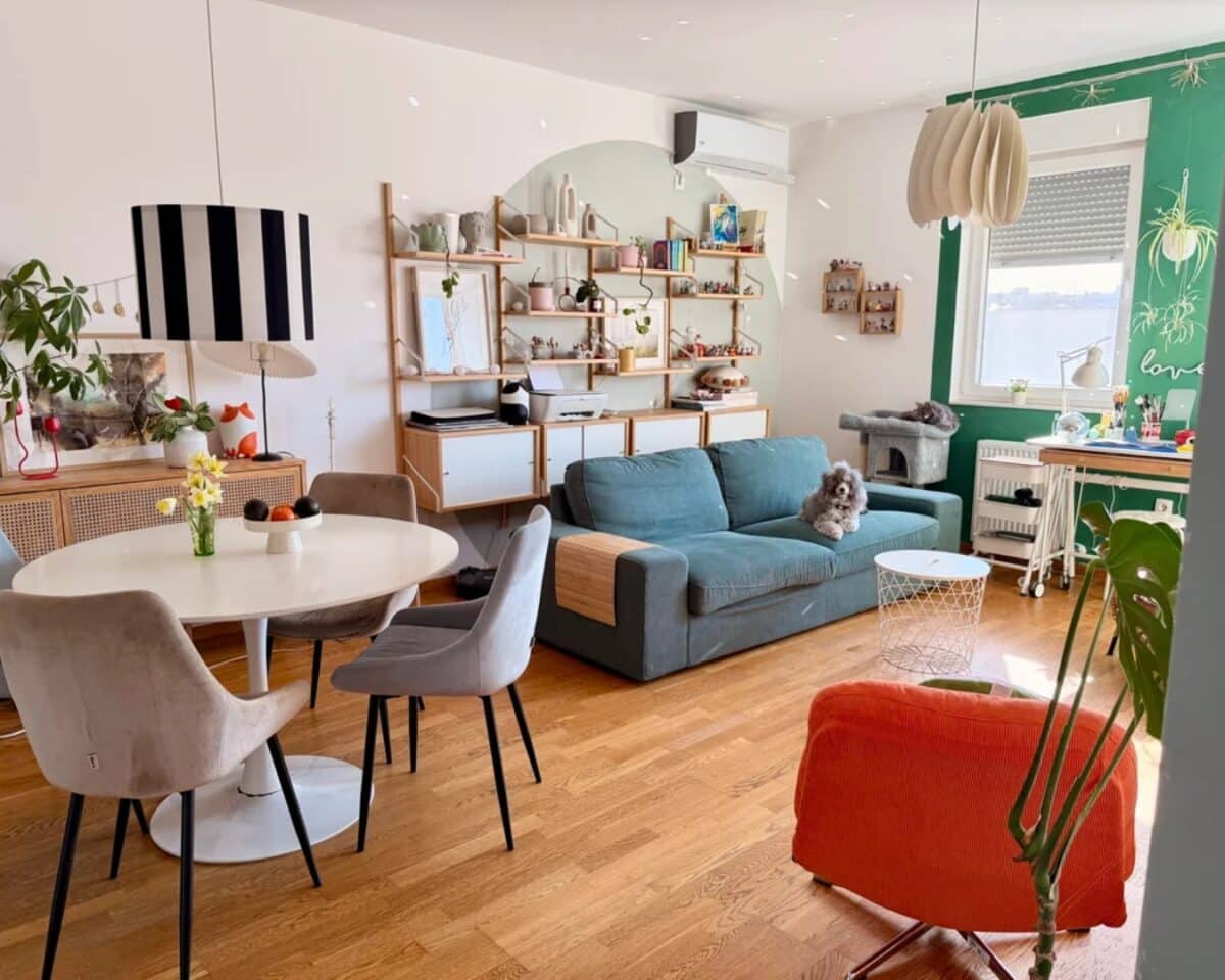 Bright open living/dining room with round white table, gray chairs, teal sofa, dog resting on the couch, and wooden shelving with decor items.