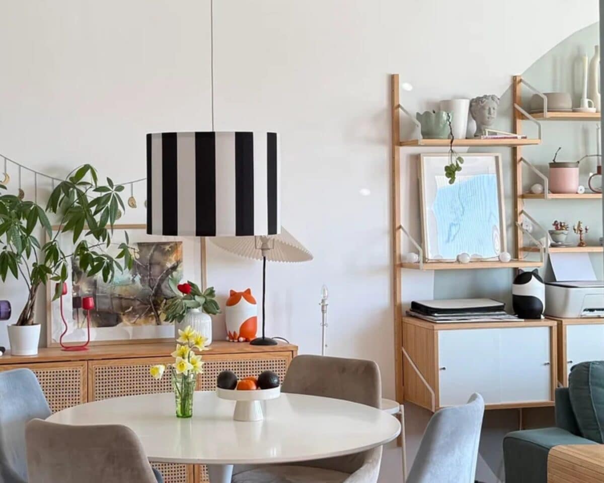 Dining area with a round white table, gray chairs, yellow flowers in a glass vase, and a tall black-and-white striped pendant lamp.