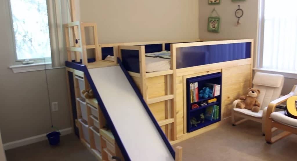 kura-bed-3-1024x557 - IKEA Hackers Wooden loft bed with a blue panel, built-in slide, and storage shelves in a child's playroom.