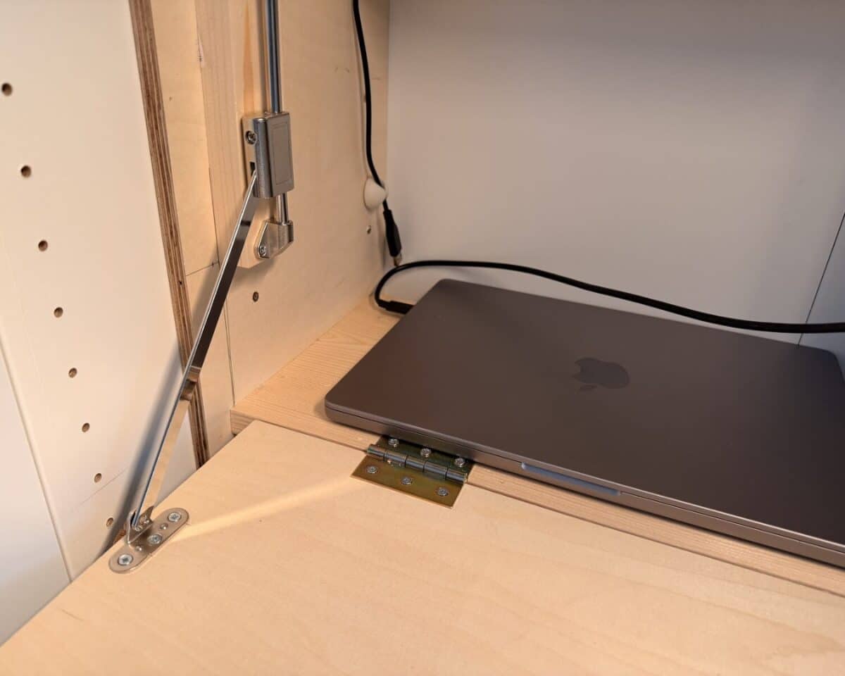 11 - IKEA Hackers Closed MacBook on a wooden shelf inside a cabinet, with metal hardware and a power cable nearby.