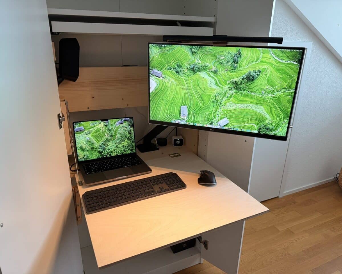 12 - IKEA Hackers IKEA PAX hideaway desk for a hidden workspace with a laptop and a large external monitor showing green terraced fields; keyboard, mouse, and a monitor arm mounted above a wooden desk lamp light.
