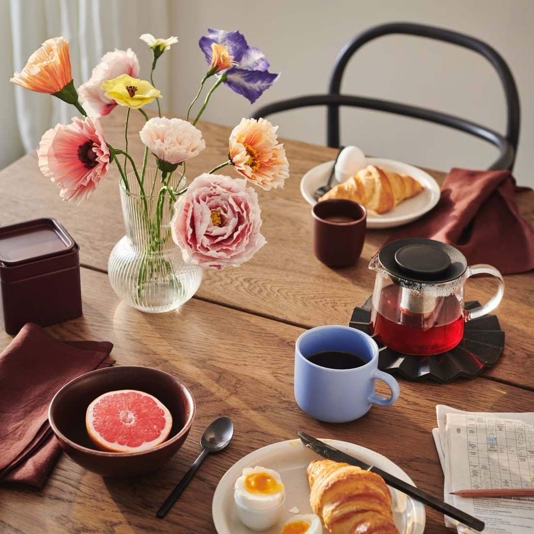 Wood dining table set for breakfast with a glass vase of colorful artificial-looking flowers, a croissant on a plate with soft-boiled eggs, half grapefruit in a brown bowl, a blue mug of coffee, a glass teapot with red tea on a hot pad, and a newspaper with a burgundy napkin nearby.