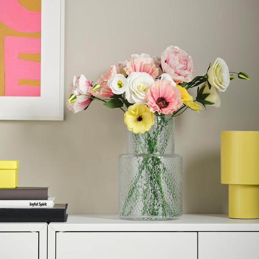 Glass vase with pink, white, and yellow flowers on a white sideboard, with yellow decorative pieces nearby and a framed artwork in the background.