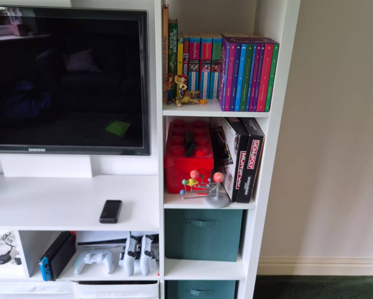 20 - IKEA Hackers White shelving unit holding a Samsung TV on the left and colorful book spines plus a Monopoly box on the right; a small solar system model and red toy bricks sit on the middle shelves, with a teal storage bin below.