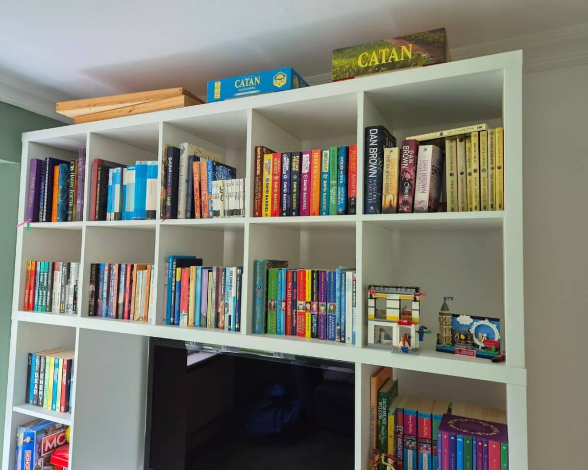 21 - IKEA Hackers White modular bookshelf filled with colorful books; a Catan box sits on the top shelf and small Lego-style sets on the right.