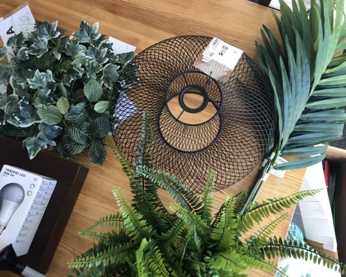 Circular black metal mesh ring on a wooden table, surrounded by artificial green plants and leaves.