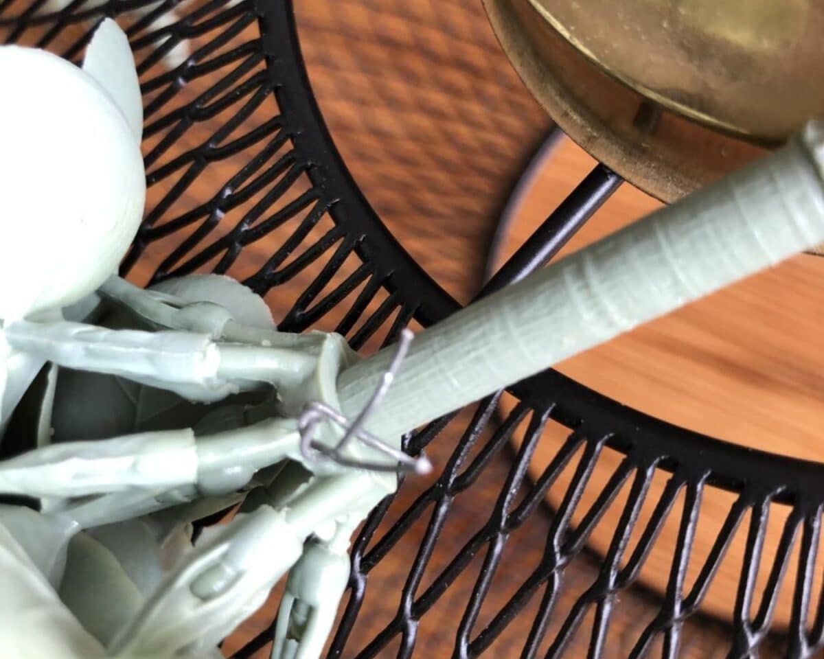 Close-up of a green plastic action figure with a small metal wire loop around its arm, next to a black wire basket surface and wood-toned background.