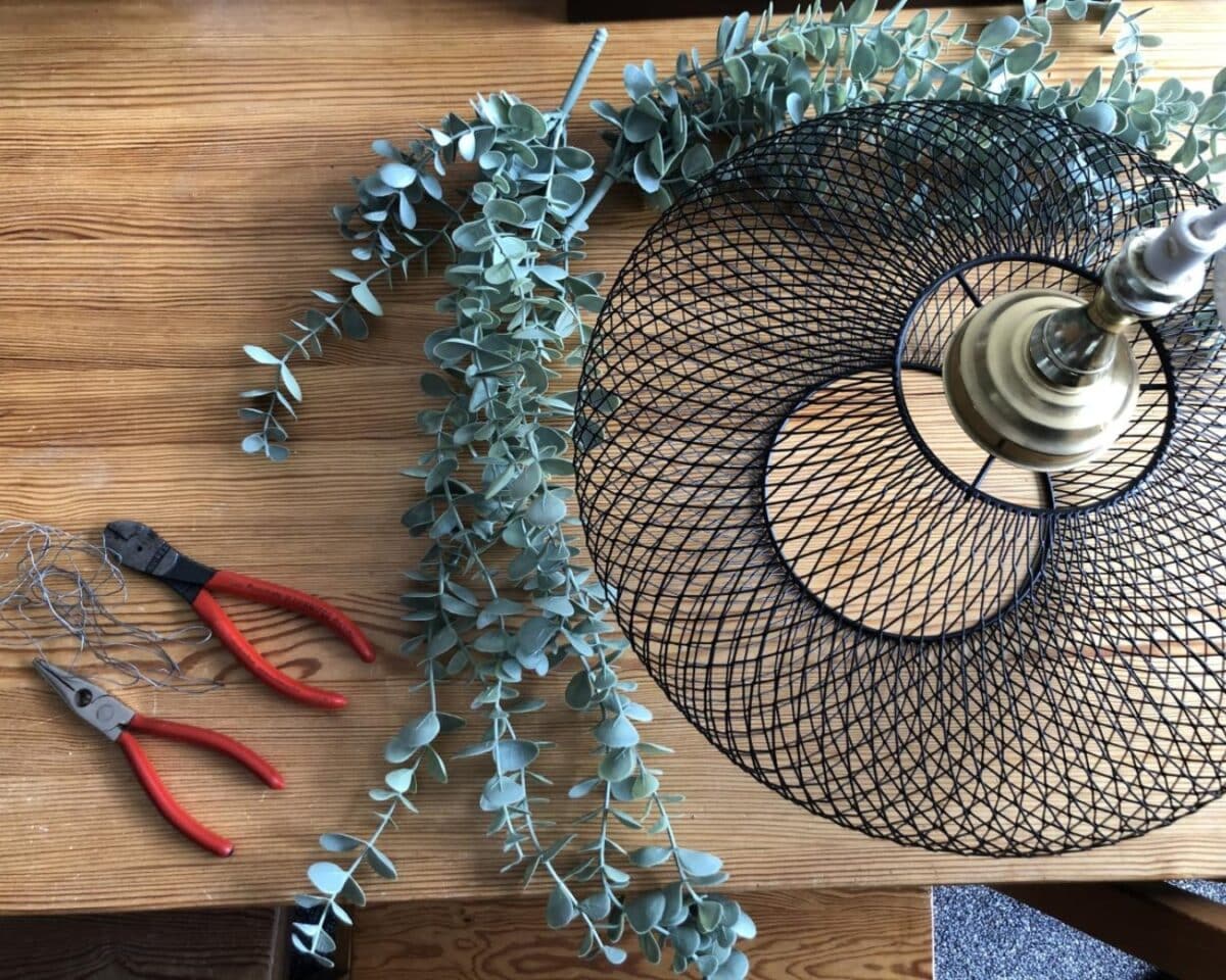 Wire mesh lampshade with a brass socket on a wooden table, next to eucalyptus garland and red-handled pliers and wire cutters—DIY setup.