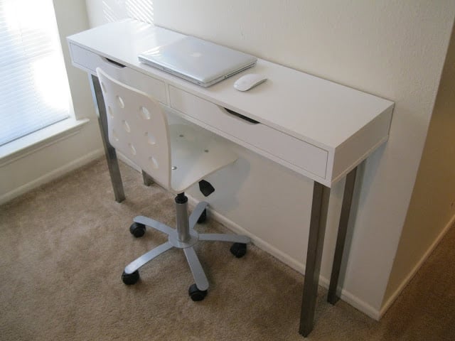 White desk with two drawers and metal legs, laptop and mouse on top, white rolling chair in a sunlit corner.