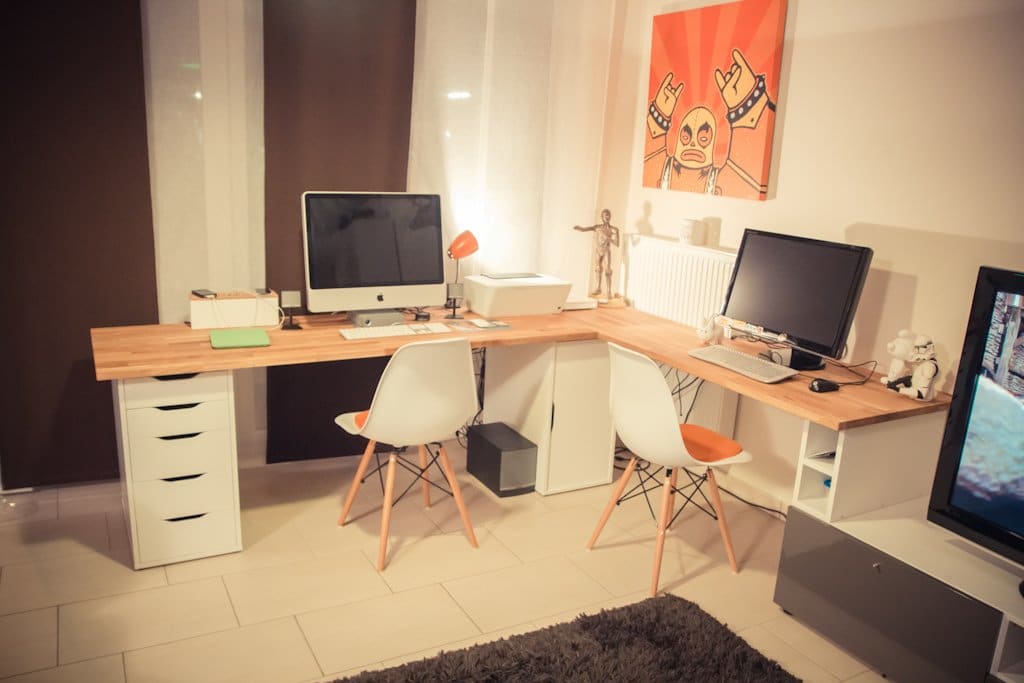 L-shaped wooden home office with two desks, an iMac and a monitor, white chairs with orange cushions, and a large red artwork on the wall.
