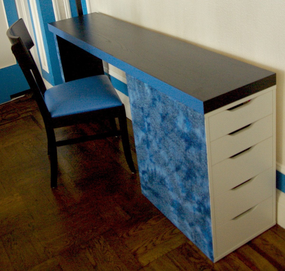 A long blue and black desk with a blue textured panel on the side and a white drawer unit on the right, plus a black chair with a blue seat pad against a striped wall.