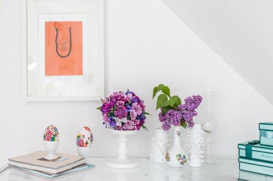 White surface with framed coral artwork on the left, a pink‑purple flower centerpiece, two small painted egg cups, and white vases holding lilac blooms, plus a stack of teal books on the right.