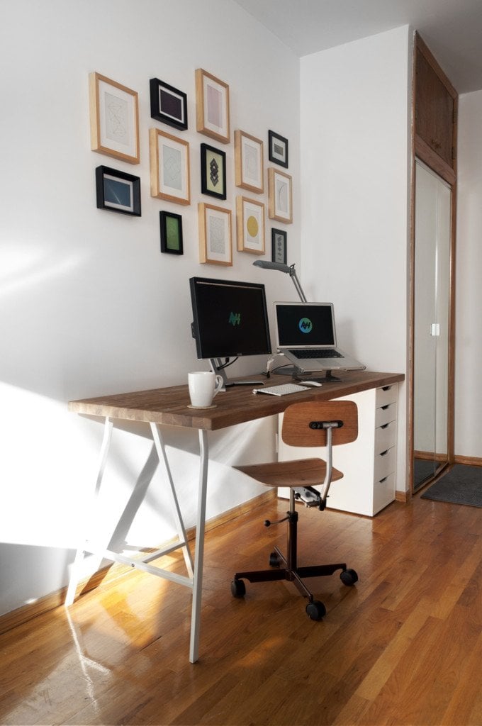 Home office desk with two monitors, a laptop, mug, and a rolling chair beside a wall of framed art.