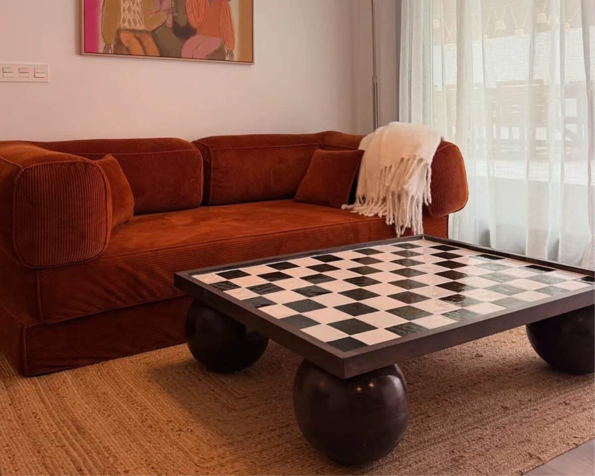 checkered-table-ikea-hack - IKEA Hackers Rust-colored sectional sofa with a cream fringed throw blanket, facing a black-and-white checkerboard coffee table on rounded black legs, in a sunlit living room with sheer white curtains.