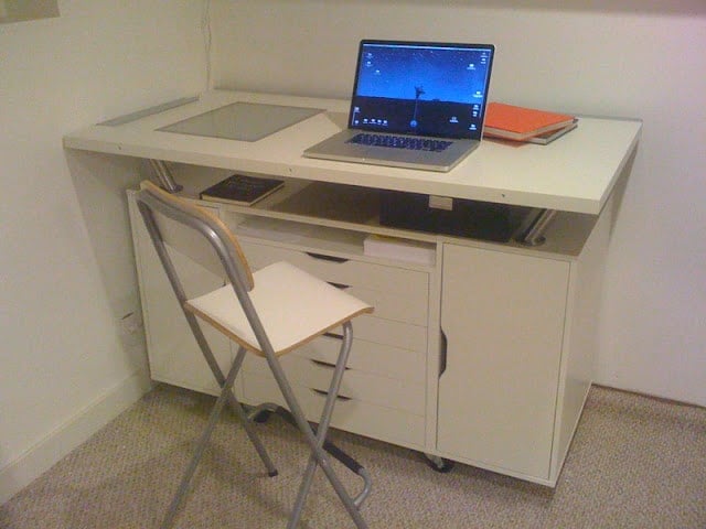 Compact white desk with a laptop on top, an orange notebook, open shelf and drawers, and a folding chair in a small home office.