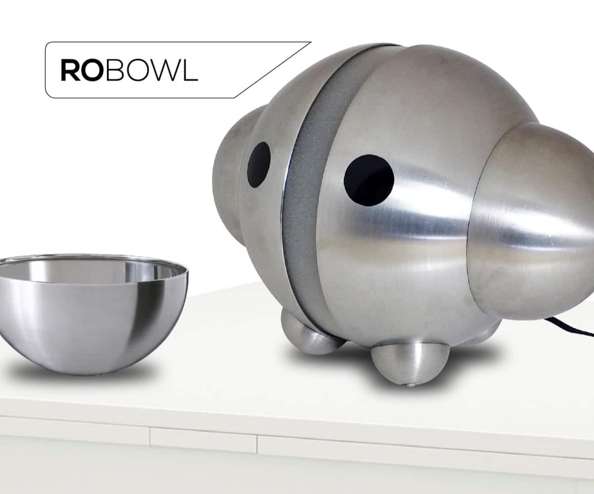 image - IKEA Hackers Robot dog-shaped stainless steel pet feeder labeled 'ROBOWL' with a separate metal feeding bowl on a white table edge.