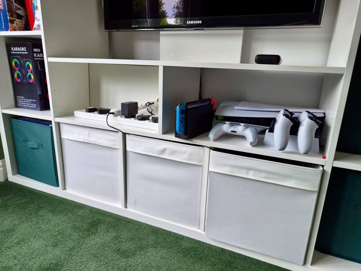 kallax lappland tv unit 4 - IKEA Hackers White modular shelf unit with a Samsung TV on top, a Nintendo Switch in its dock with blue and red Joy-Con controllers, plus two white controllers on the right, and fabric storage bins below on green carpeted flooring.