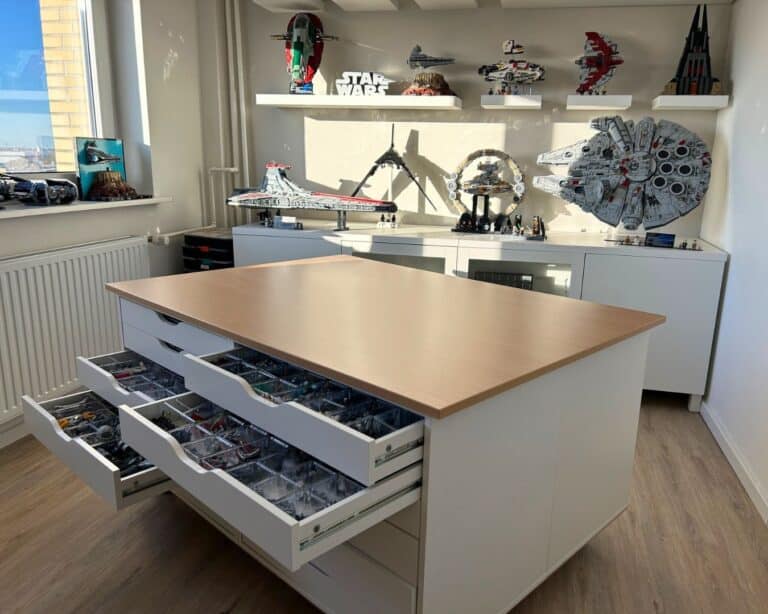 Bright workshop room with a central island worktable and several drawers open to reveal organized LEGO bricks; wall shelves display Star Wars LEGO ships and models, including a large Millennium Falcon-like craft.