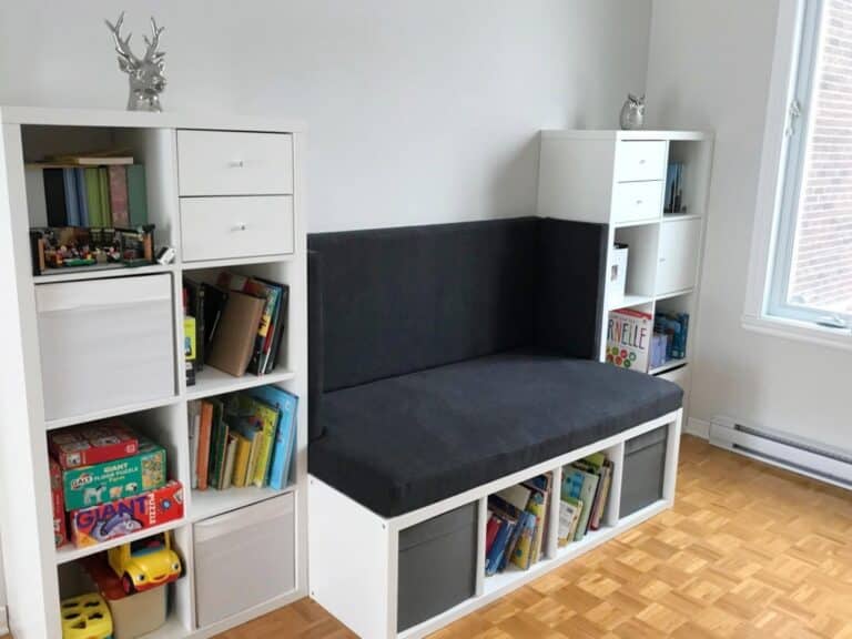 reading-nook-bench-kallax-hack-768x576 - IKEA Hackers White modular storage units form a seating nook with a gray cushion, books, and toys on shelves in a bright room.