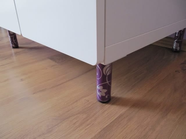 Close-up of a purple patterned furniture leg with decorative wrap under a white cabinet on a wooden floor.