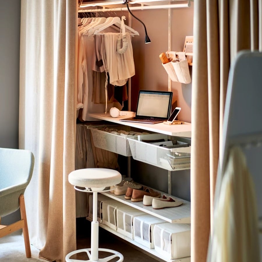 19 - IKEA Hackers Laptop on a white desk inside a wardrobe area, with phone, sewing organizers, and shoes on the lower shelf, surrounded by hanging clothes and curtains.