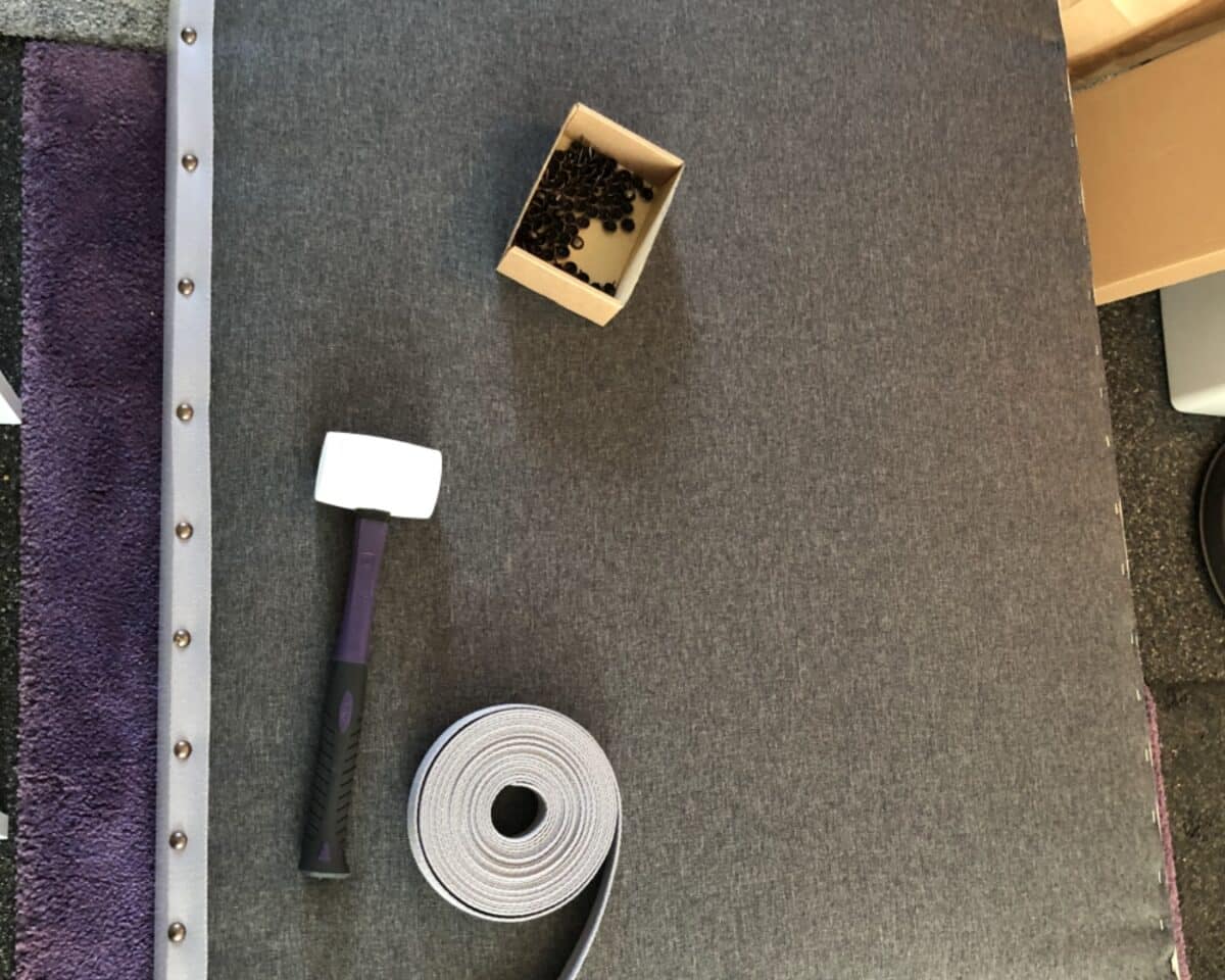 Hammer with white head and purple handle, a box of small black nails, and a roll of gray packing strap on a work surface.