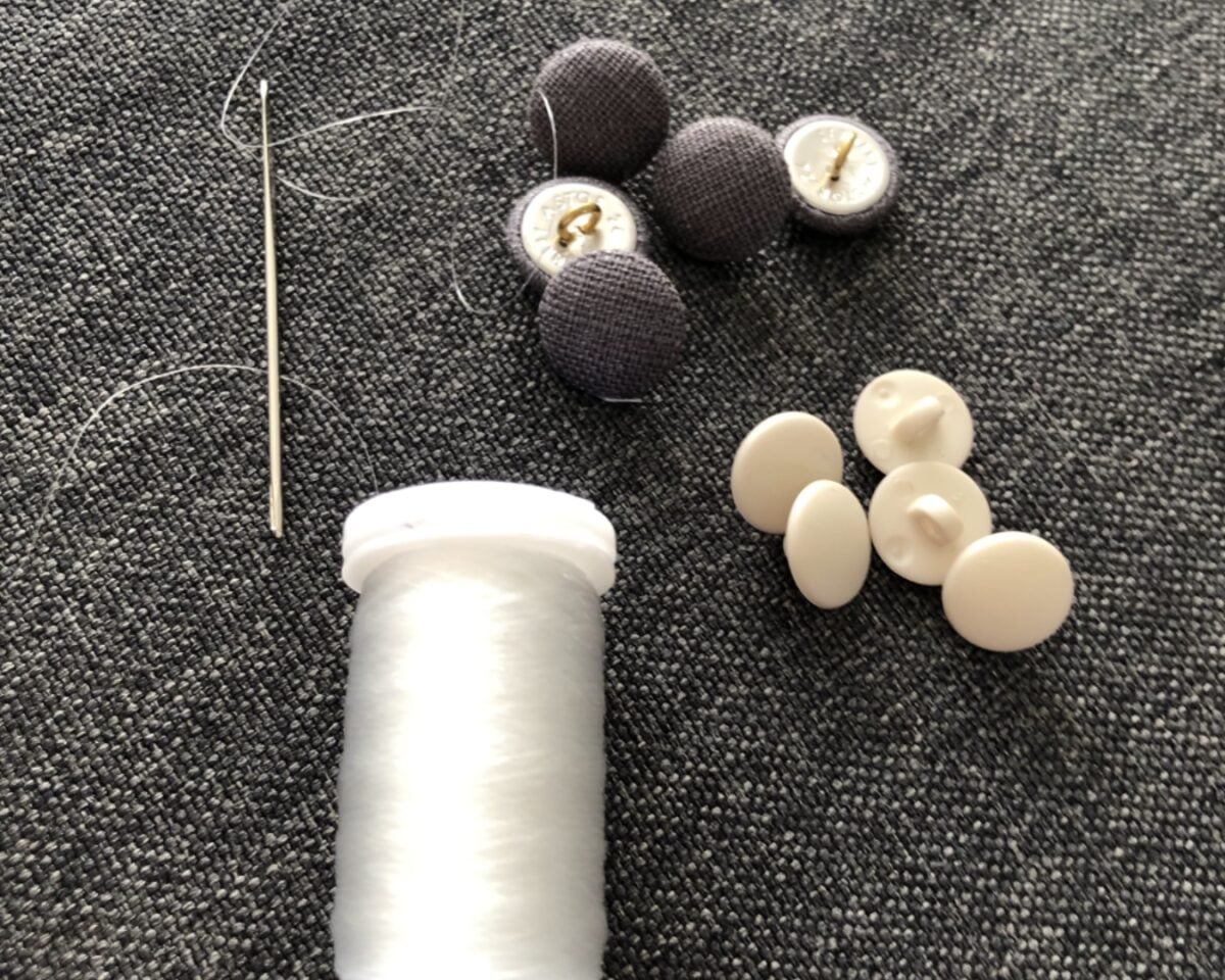 Sewing supplies: white thread spool with needle, black fabric-covered buttons, and white disk buttons on dark fabric.