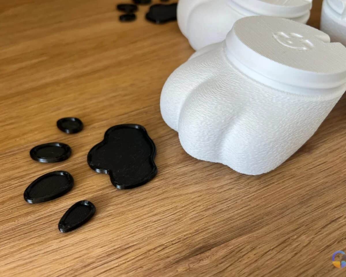White textured 3D-printed paws with a circular lid on a wooden surface, surrounded by black plastic grippers