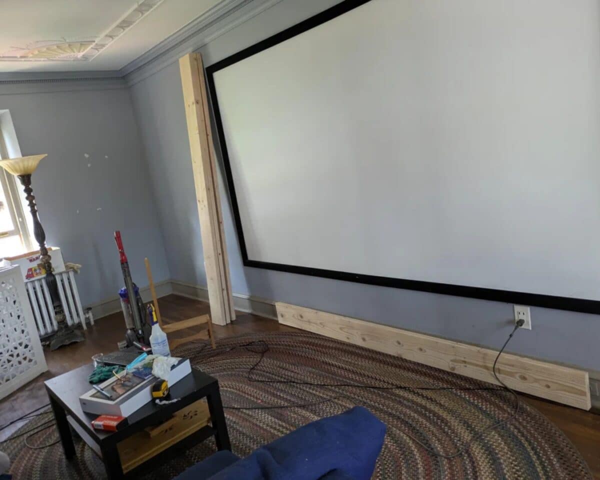 Home theater setup in progress: a large pull-down projector screen mounted on a blue wall, with wooden supports, a vacuum, and a cluttered coffee table nearby.