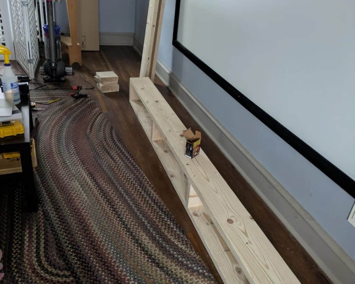 Indoor workshop scene with a long pine board resting on a two-tier wooden bench along a blue wall, plus tools and a patterned rug nearby.