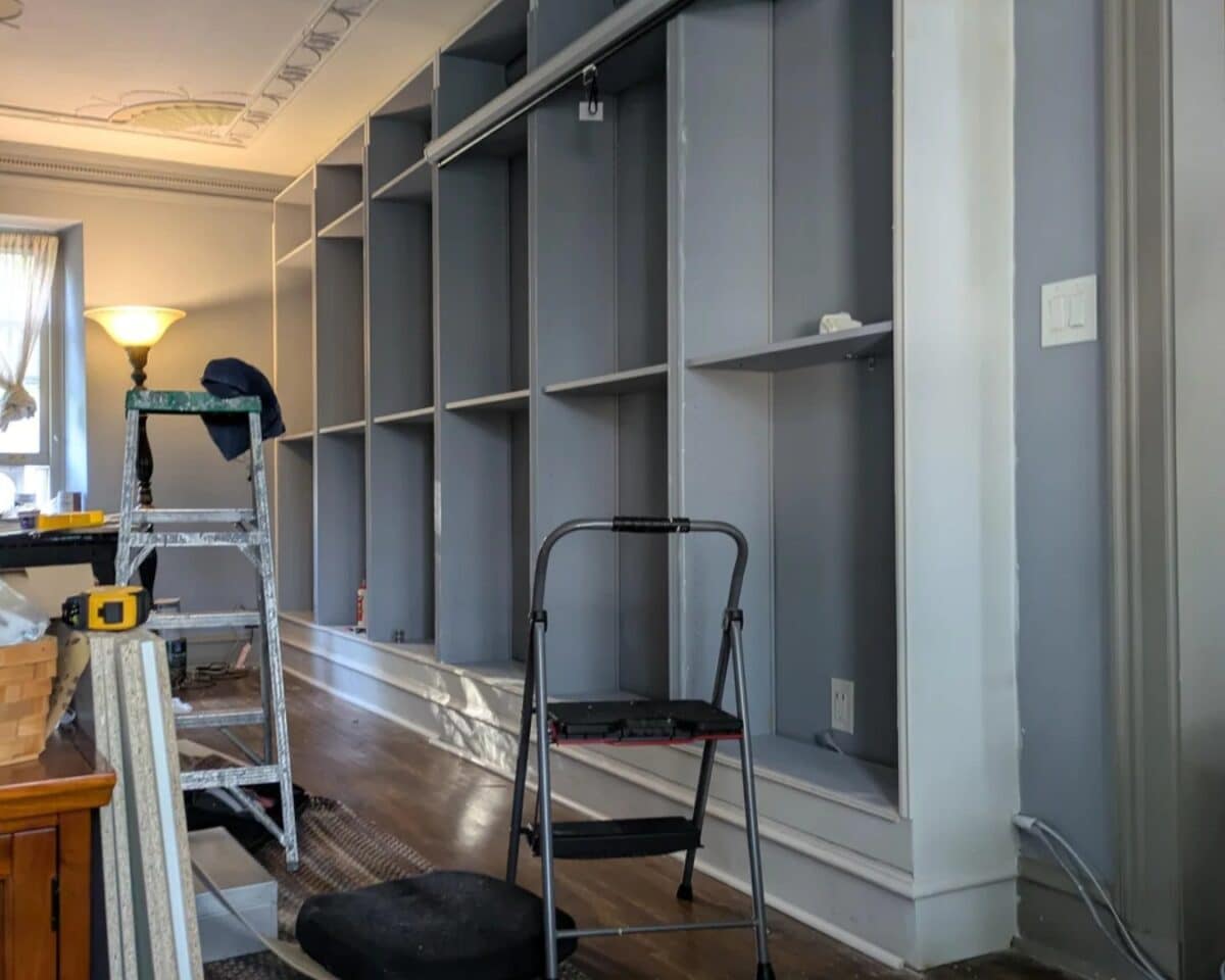 Construction in progress: tall gray built-in shelves along the wall with a ladder and tools nearby.",